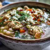 A hearty Greek Chicken & Pearl Couscous Soup garnished with lemon wedges, feta, and dill.