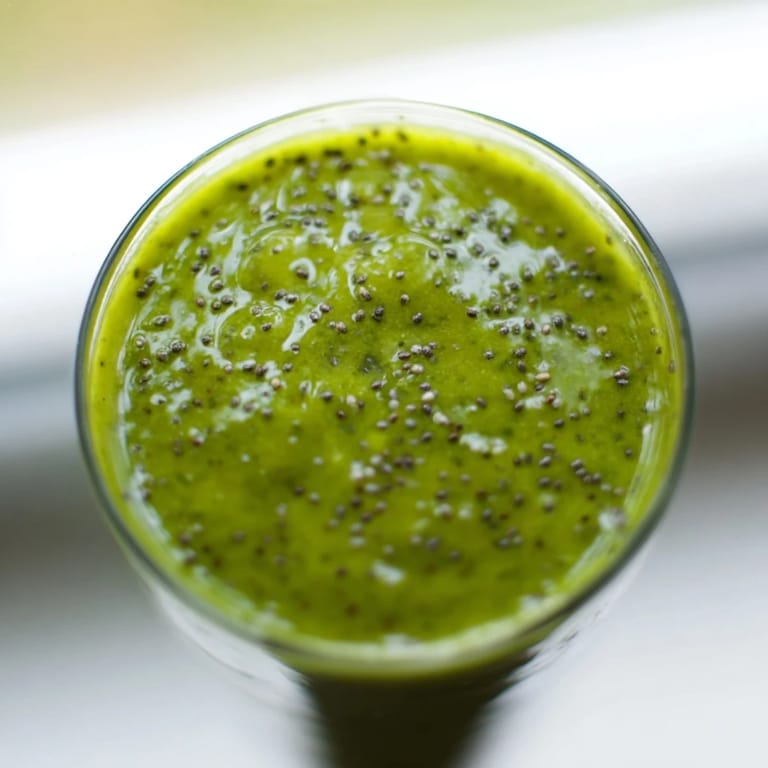 Glass of green detox smoothie beside sliced apple, lemon wedge, and chia seeds, served over ice for a refreshing morning start.